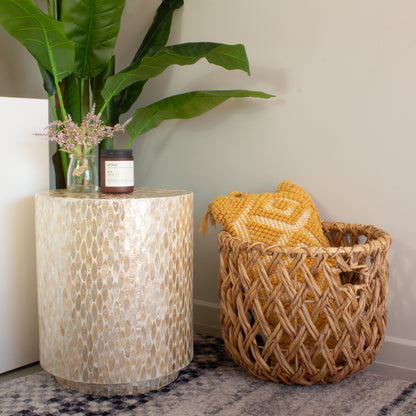 Ely 40cm Mother of Pearl Inlay Side Table in Beige, featuring a stunning mosaic surface, is adorned with a small glass jar of pink blooms and a dark-glass candle, complemented by a mustard-yellow throw in a wicker basket nearby.