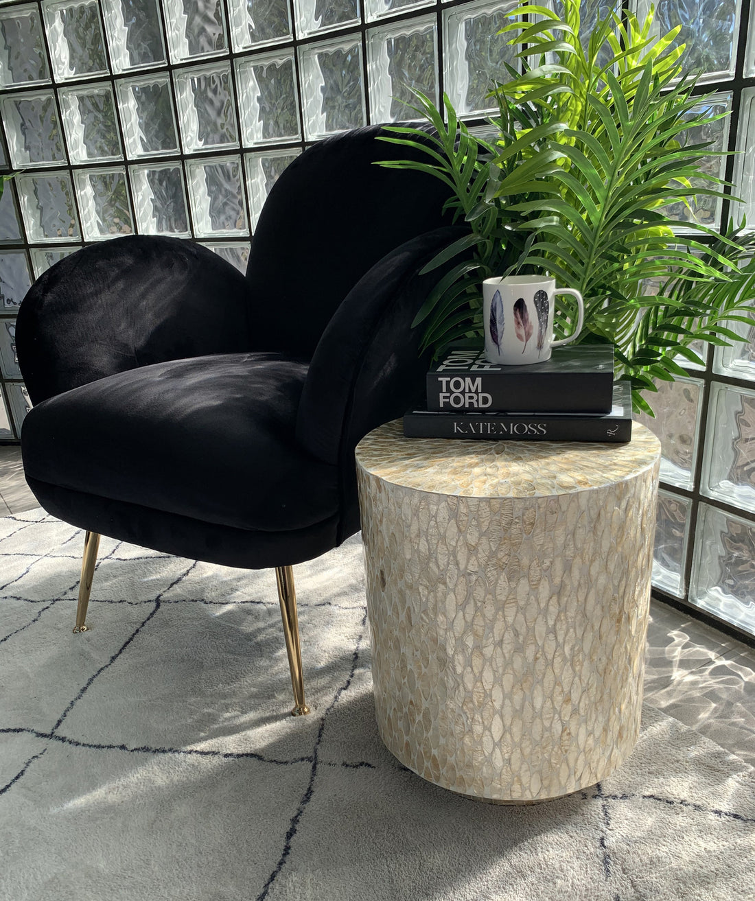 The Ely 40cm Mother of Pearl Inlay Side Table in Beige features a cylindrical design with a stunning mother-of-pearl mosaic surface, complemented by elegant gold metallic feet, beside a cozy black velvet armchair.