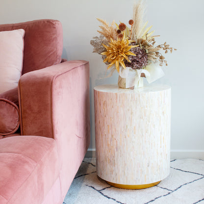Ely 40cm Mother of Pearl Inlay Side Table in Cream, featuring a luminous mosaic surface and gold base, paired with a dried floral arrangement, beside a plush blush-pink velvet sofa.