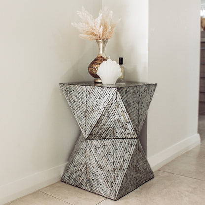 Enki 35cm Mother of Pearl Inlay Side Table in Beige & Black, featuring a geometric hourglass shape with a silvery mosaic finish, topped with a metallic vase, candle, and perfume bottle in a tiled corner.