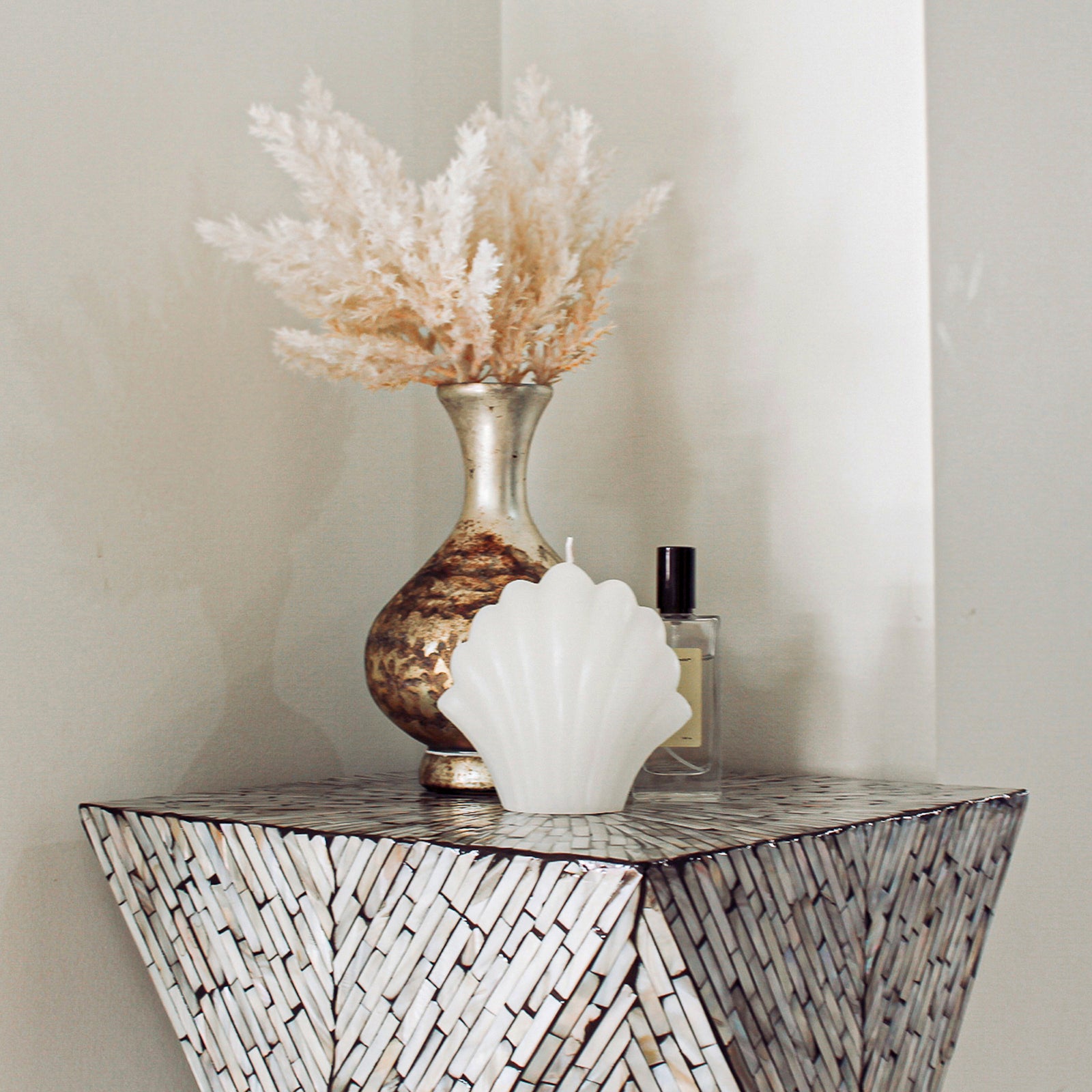 Enki 35cm Mother of Pearl Inlay Side Table in Beige & Black with mosaic detailing, showcasing a metallic vase with pampas grass, scallop shell candle, and glass perfume bottle against a neutral backdrop.