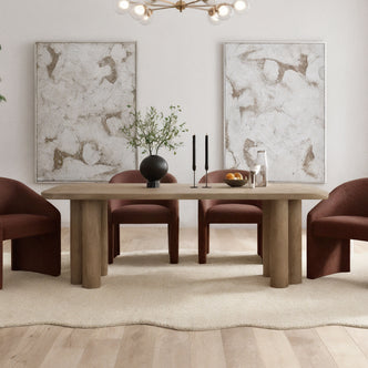 Faunus 200cm 8 Seater Dining Table in Natural with chunky cylindrical legs, centered on a pale rug, surrounded by rust-upholstered chairs, adorned with a black vase, candles, and a fruit bowl.