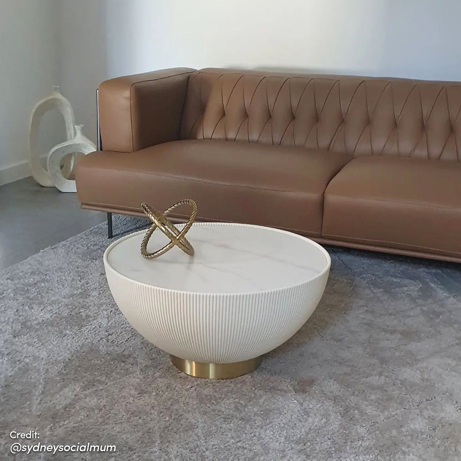 Glalie 70cm Sintered Stone Coffee Table Cream White with fluted base and marble-look top, accented by a brass pedestal, on a plush gray rug, featuring a decorative brass sculpture.
