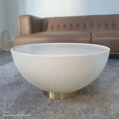 Glalie 70cm Sintered Stone Coffee Table Cream White with a low, ribbed half-sphere form, marble-look top, and brushed brass plinth base, placed in a modern living-room setting.