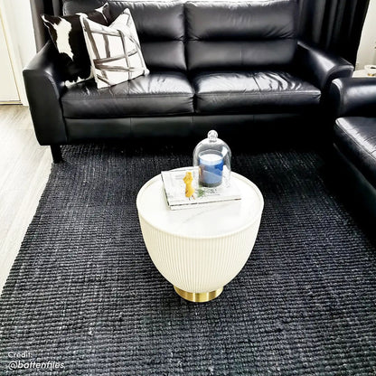 Glalie 47cm Sintered Stone Coffee Table in Cream White, a round fluted drum design on a low brass base, topped with books and a yellow bunny figurine under a glass cloche.