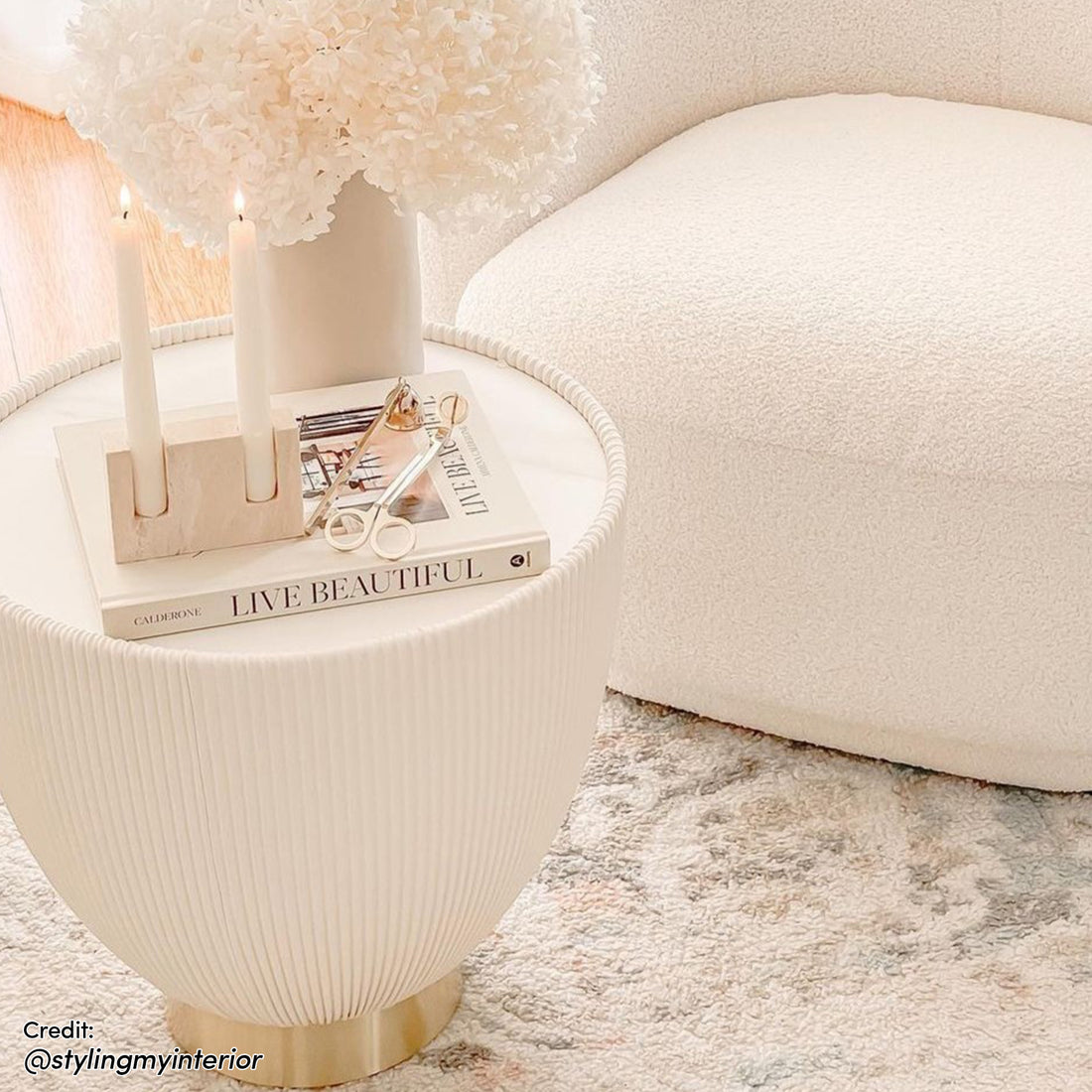 Glalie 47cm Sintered Stone Coffee Table features a ribbed white surface and brass base, topped with a book, marble candleholder, and hydrangeas, complemented by a boucle chair in a cozy setting.