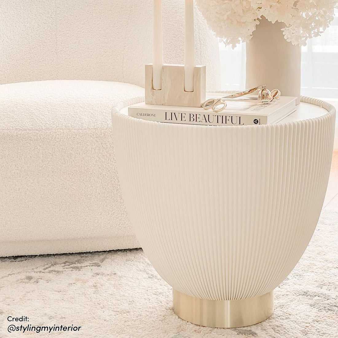 Glalie 47cm Sintered Stone Coffee Table in Cream White with a fluted shell design and polished brass base, topped with hydrangeas, a candle holder, and a book.