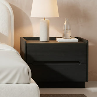 Glorinda 60cm Scratch Resistant Bedside Table in Black featuring two drawers, sleek design, and a lamp, books, and glass carafe on top, next to a white-upholstered bed.