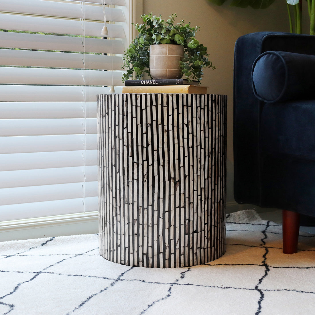Ivo Mother of Pearl Inlay Side Table in Ivory & Black with a vertical strip mosaic pattern, topped with books and a plant, beside a dark blue sofa and light area rug.
