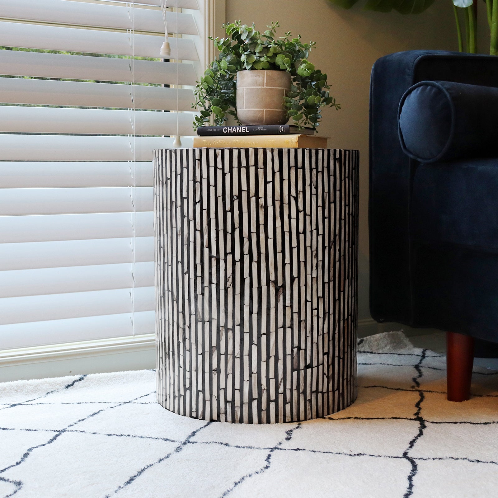 Ivo Mother of Pearl Inlay Side Table in Ivory & Black with a vertical strip mosaic pattern, topped with books and a plant, beside a dark blue sofa and light area rug.