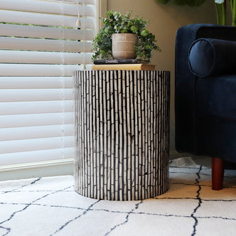Ivo Mother of Pearl Inlay Side Table in Ivory & Black with a vertical strip mosaic pattern, topped with books and a plant, beside a dark blue sofa and light area rug.