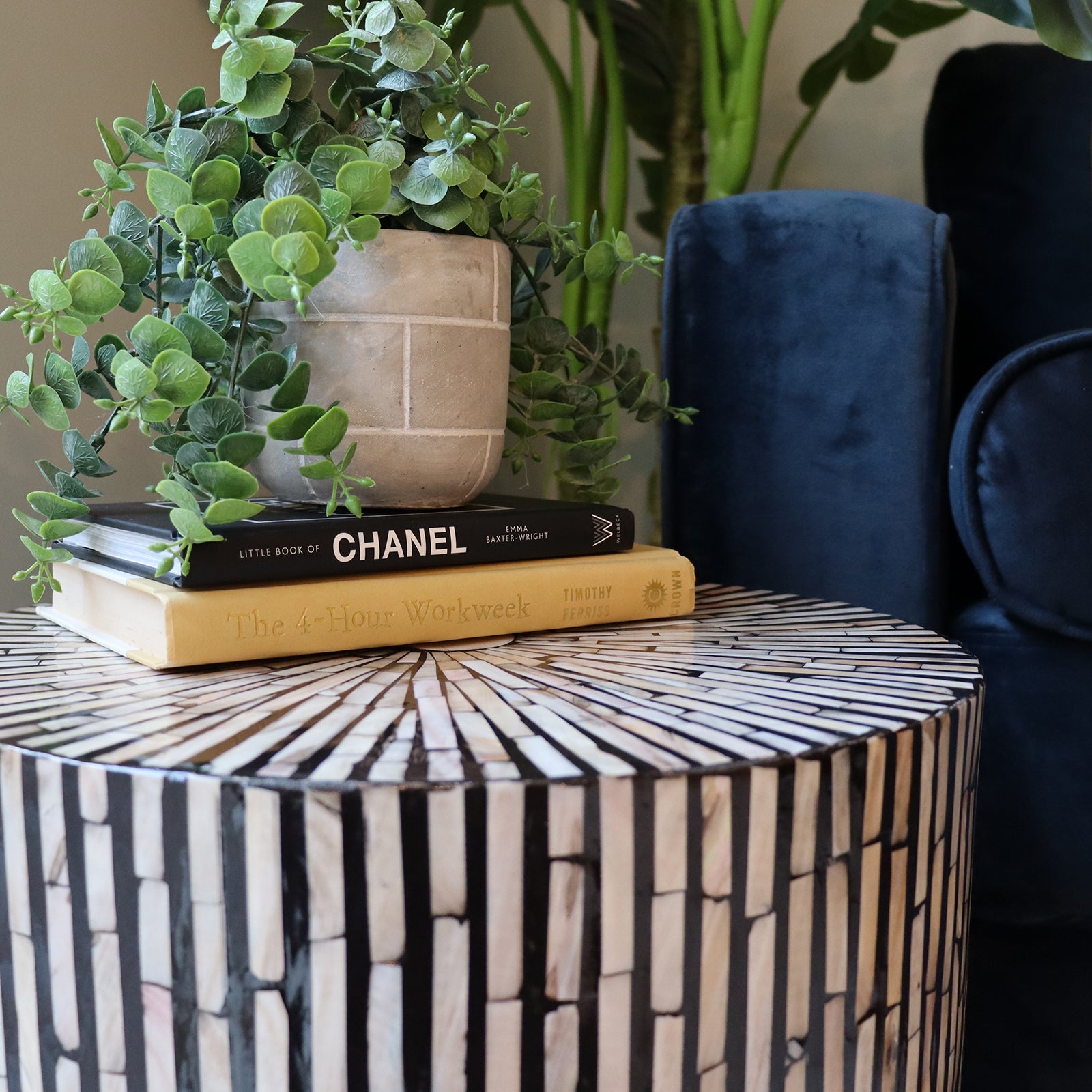 The Ivo Mother of Pearl Inlay Side Table in Ivory & Black features a mosaic pattern, topped with books and a planter, alongside a deep-blue chair, enhancing any living space.