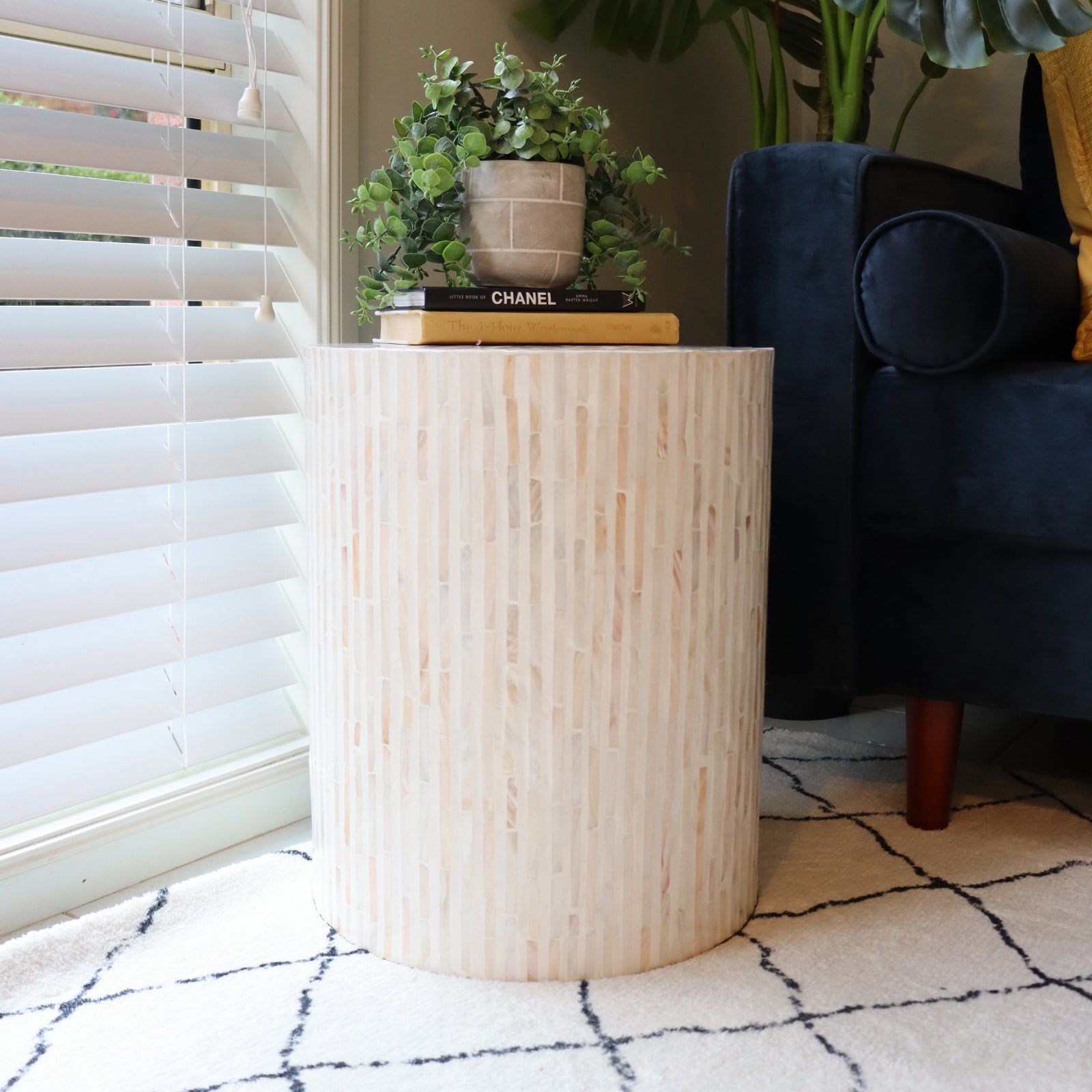 Ivo Mother of Pearl Inlay Side Table in Cream features a pale mosaic veneer, topped with books and a faux plant. Positioned on a grid-patterned rug beside a dark sofa.