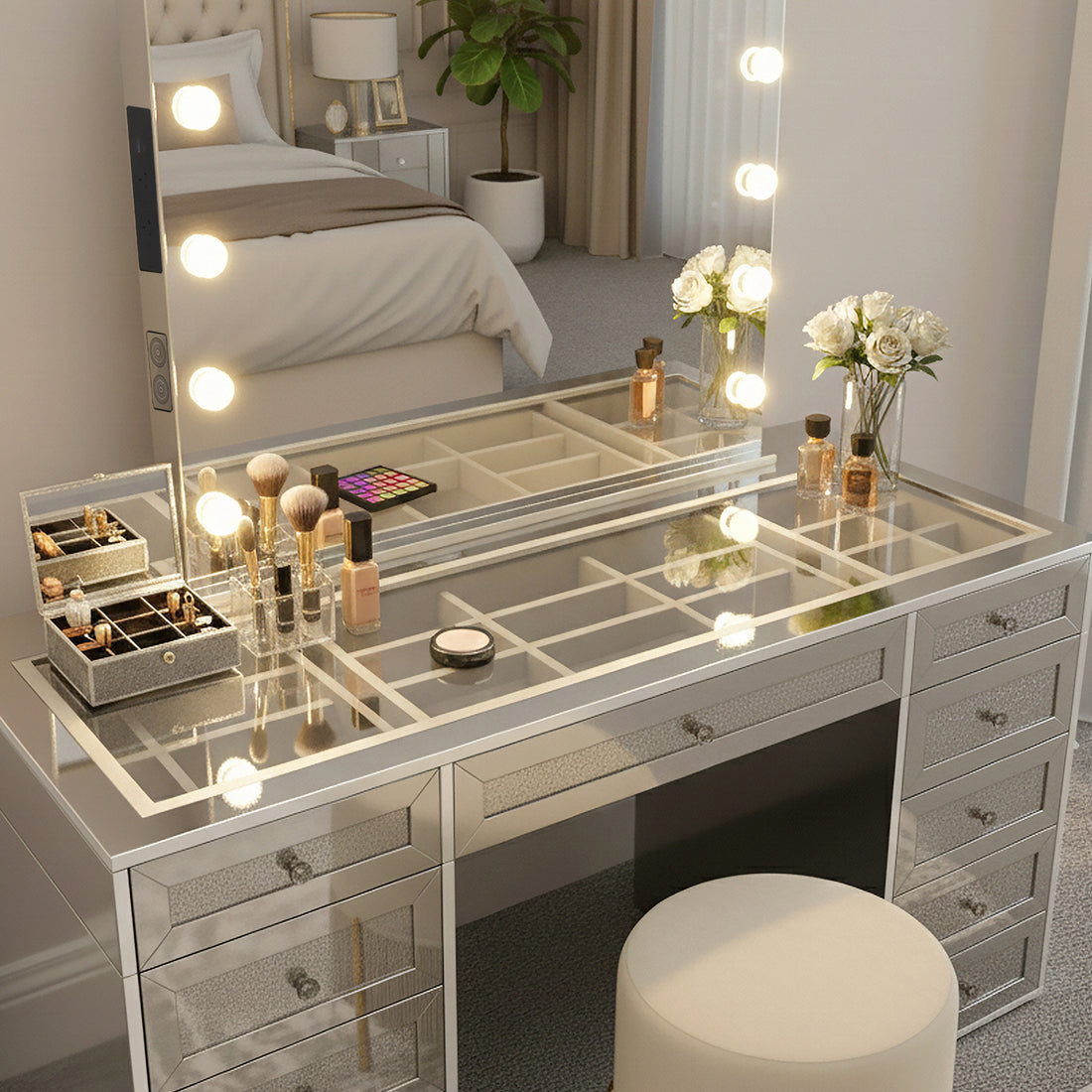 Ivy Diamond Hollywood Vanity Table in Silver, featuring a mirrored design with LED bulb-framed mirror, glass top, organized makeup display, white roses, mirrored drawers, and an upholstered stool for a luxurious setup.