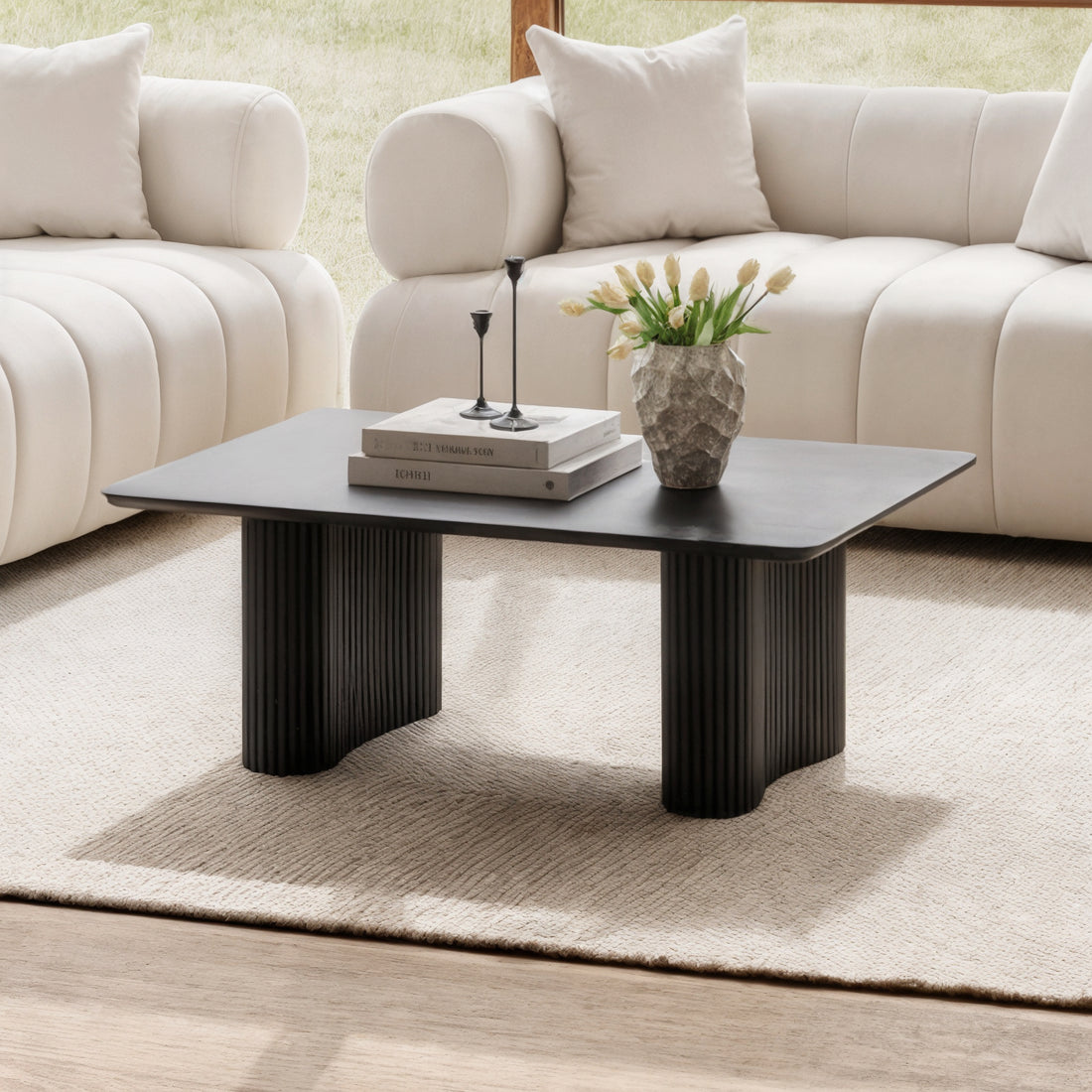 Joyce 120cm Coffee Table with Round Fluted Legs in Black, featuring a sleek rectangular top styled with books, candleholders, and a vase, set on a woven rug between cream sofas.