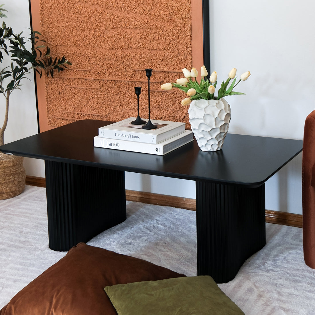 Joyce 120cm Coffee Table with Round Fluted Legs in Black on a light-gray rug, adorned with books, matte-black candle holders, and a white ceramic vase with tulips, surrounded by cushions and a planter.