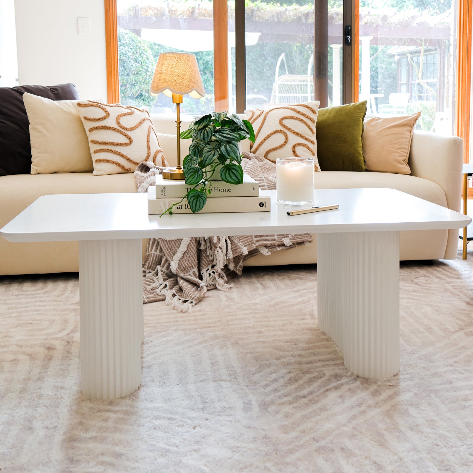 Joyce 120cm Coffee Table with Round Fluted Legs in White, adorned with books, a potted plant, a lit candle, and a pen, enhances a bright, airy living room setting.