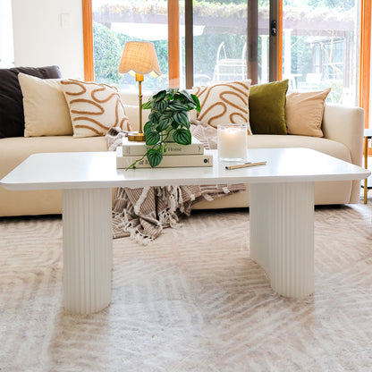 Joyce 120cm Coffee Table with Round Fluted Legs in White, adorned with books, a potted plant, a lit candle, and a pen, enhances a bright, airy living room setting.
