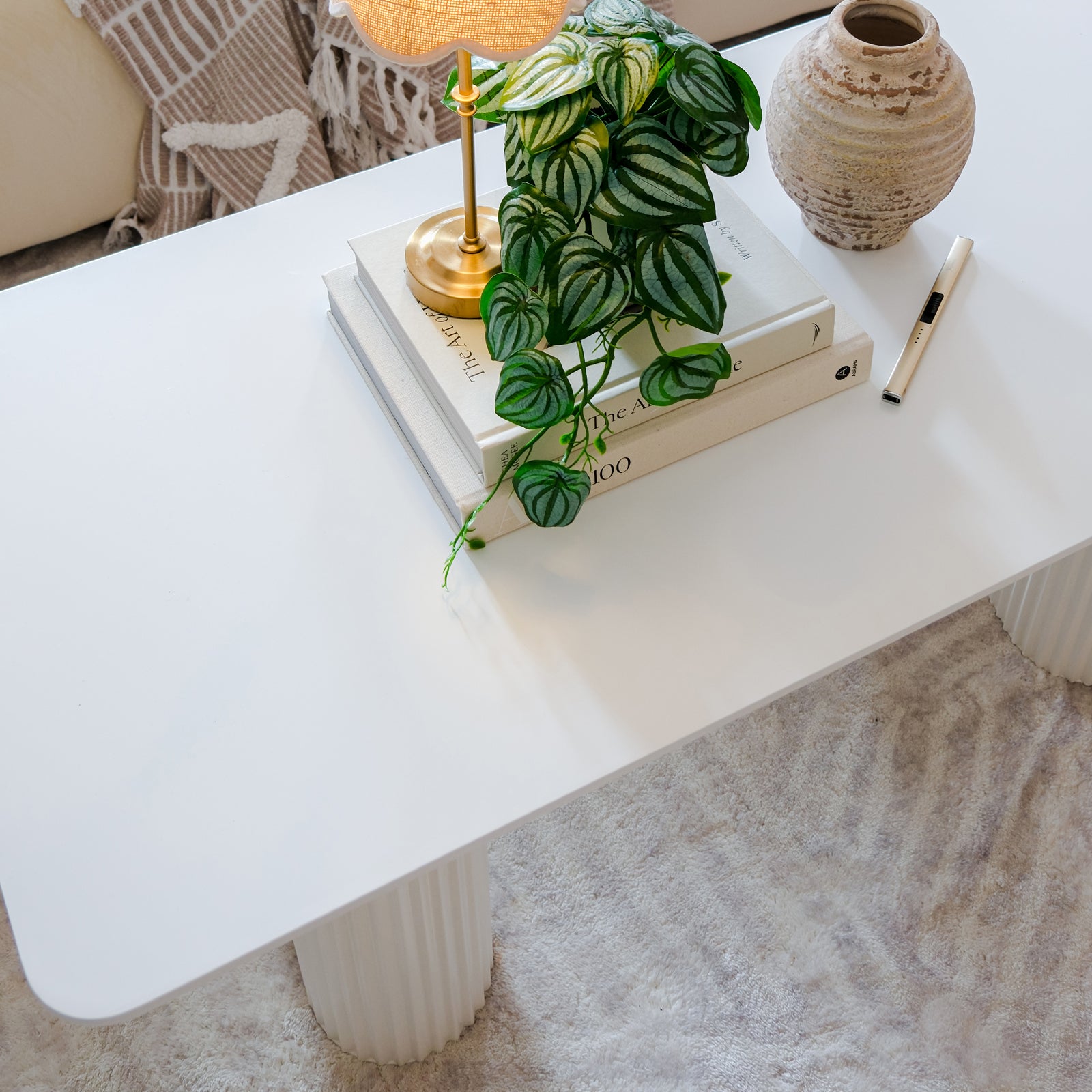 Joyce 120cm Coffee Table with Round Fluted Legs in White, featuring a minimalist design with books, a brass desk lamp, a trailing houseplant, ceramic vase, and a slim object, on a neutral rug.