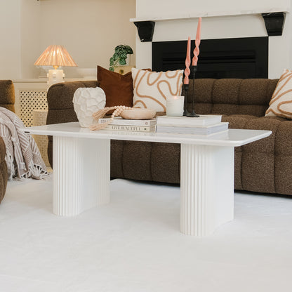 Joyce 120cm Coffee Table with Round Fluted Legs in White, styled with books, a vase, a bowl with beads, a lit candle, and pink candles, set in a cozy living-room vignette.