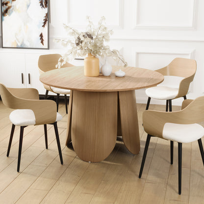 Kalyani 120cm Wooden Round Dining Table with Veneer Top Oak, featuring a sculptural petal-like base, surrounded by five light-wood chairs with white upholstered seats and slender black legs.