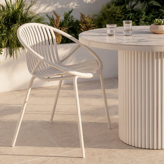 Kosmos Outdoor Round Dining Chair Set of 2 in White, featuring a modern slatted design with curved arms and legs, paired with a round table holding glasses and a succulent, set on a stone patio.