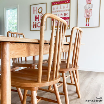 Leigh Solid Oak High-back Dining Chair Natural Set of 2 in a minimalist dining space, showcasing spindle-back design and sturdy oak build around a light-wood table, accented by holiday-themed wall prints.