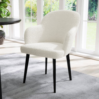 Lennon Boucle Dining Chair Set of 2 in Cream White, featuring soft boucle fabric and slender black legs, placed on a gray rug near large windows, with a table and plant nearby.
