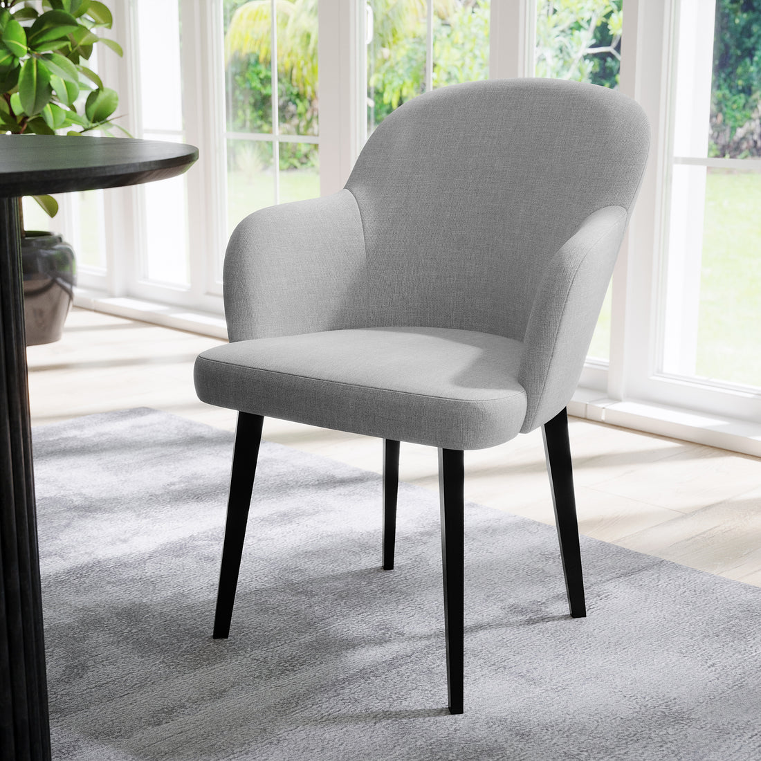 Lennon Linen Dining Chair Set of 2 in Light Grey features modern upholstered design with slim black legs, set on a soft rug beside a round table, in front of sunlit windows.