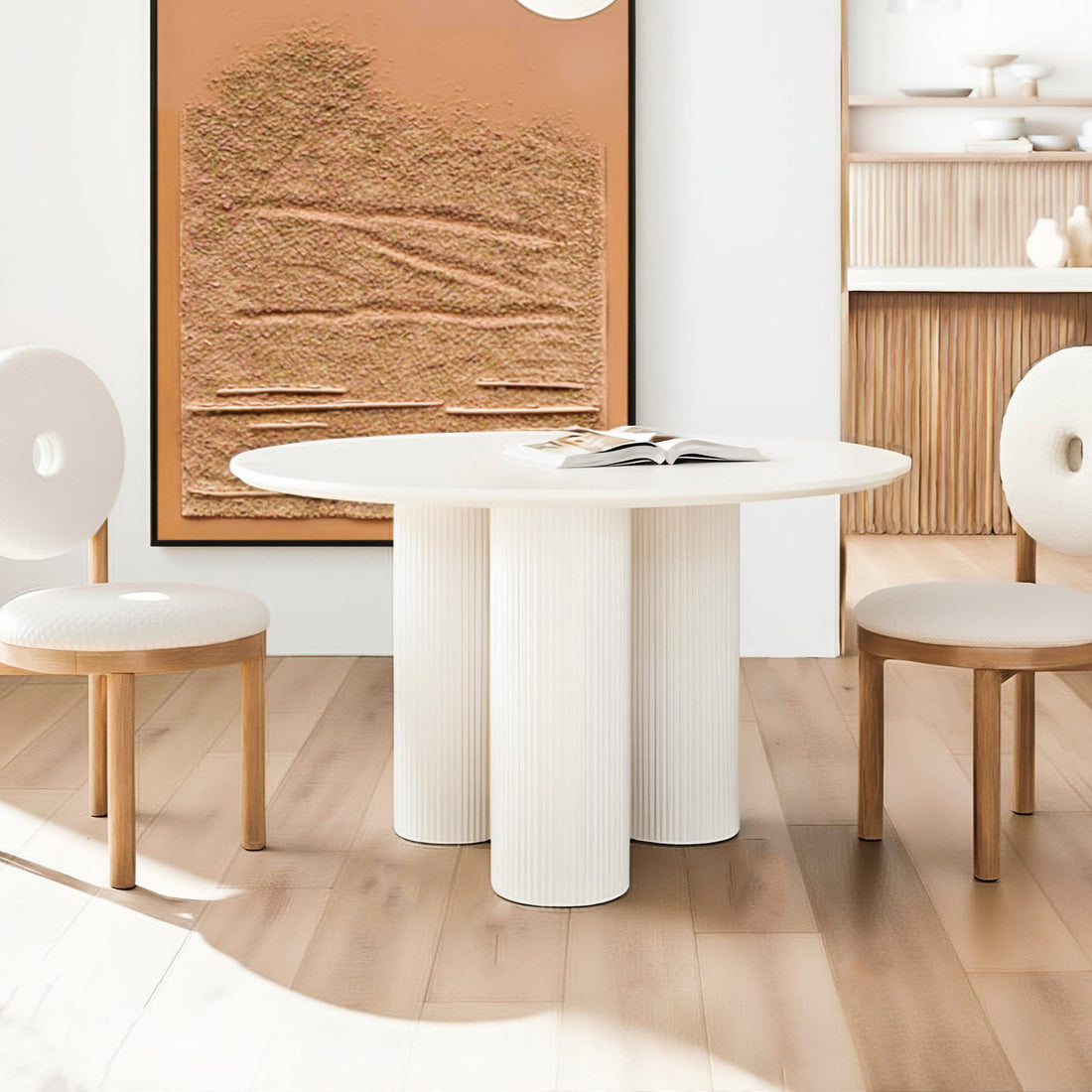 Lucina 4 Seater Fluted Round Dining Table in White with sculptural pillar legs, set in a minimalist nook alongside matching side-chairs and a rust-colored wall panel, creating a modern, elegant dining space.