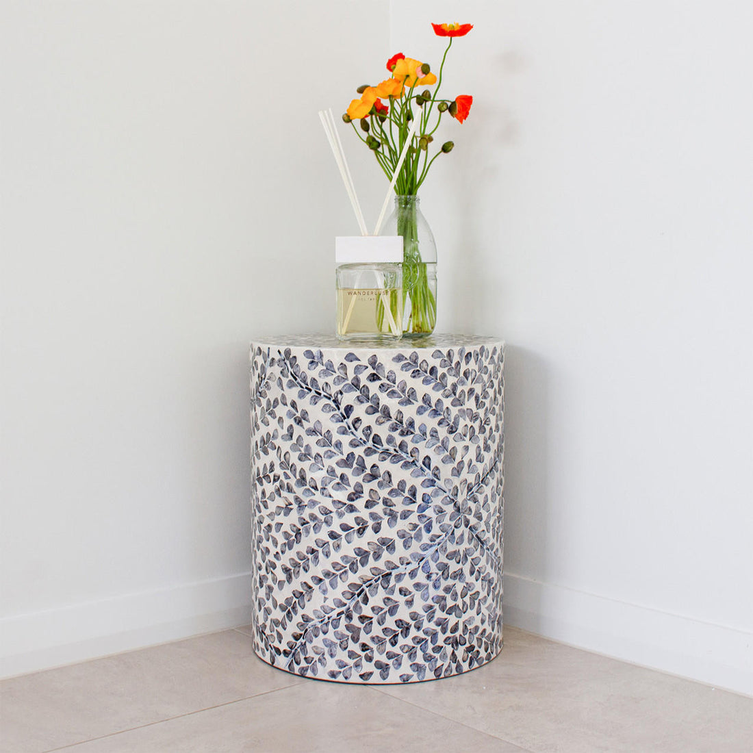 Lulu Floral Mother of Pearl Inlay Side Table in Blue, showcasing a white and gray leaf-patterned inlay topped with a glass bottle of orange flowers and a reed diffuser, set against pale walls.