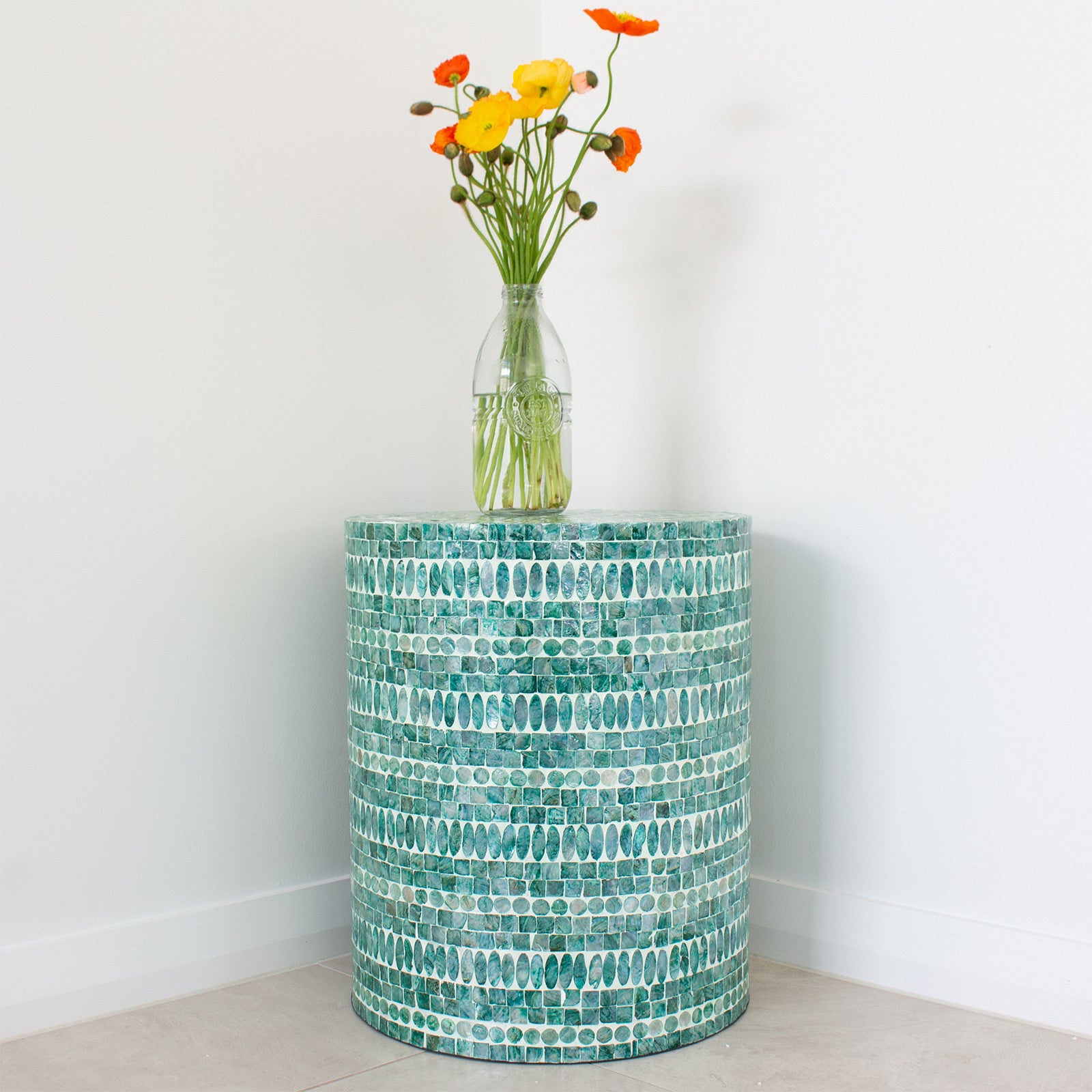 Lulu Mother of Pearl Inlay Side Table in Jade Green, round with green-blue mosaic tiles, topped with a clear glass vase holding yellow and orange flowers against a plain white wall.