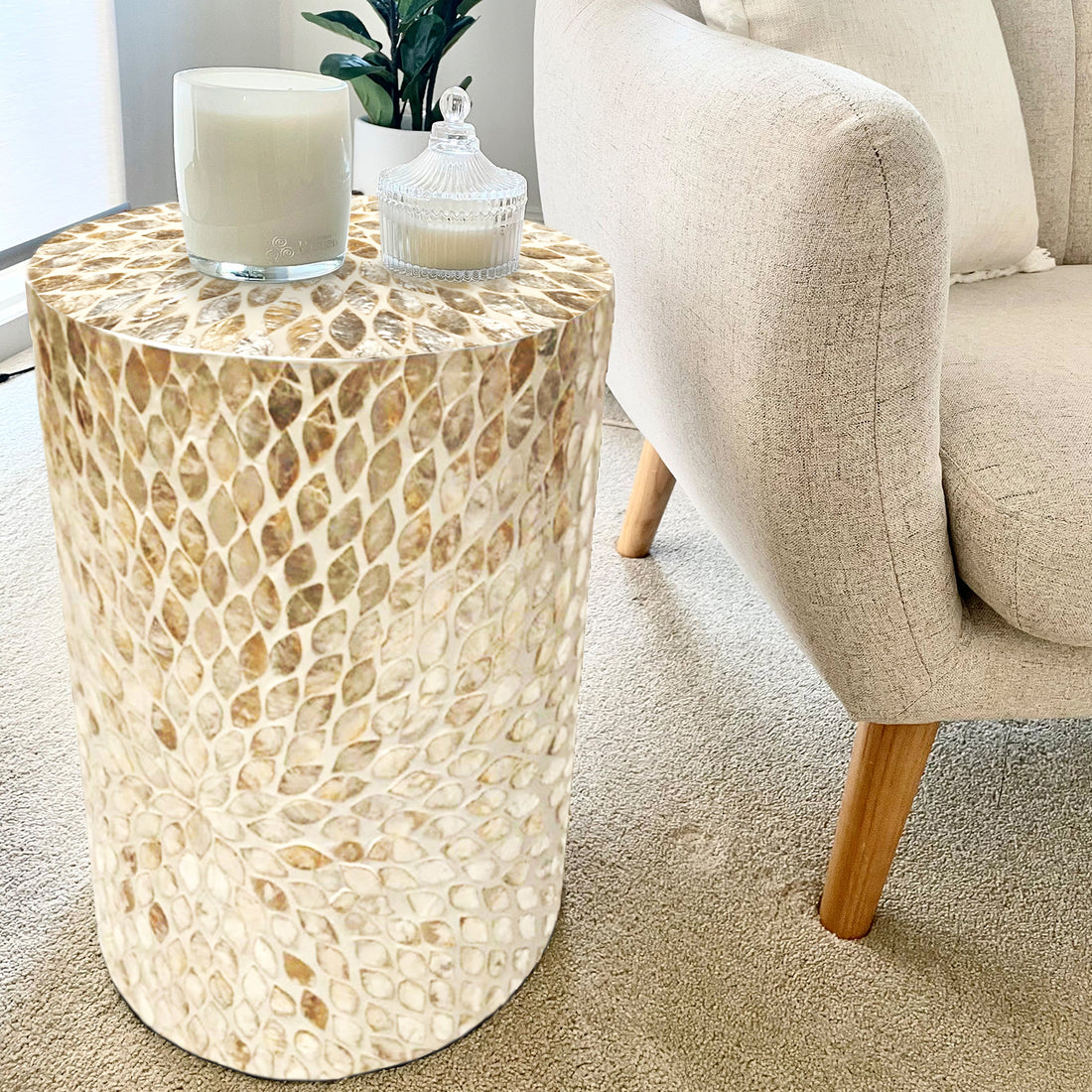 Lulu Mother of Pearl Inlay Side Table in Sand, featuring a light mosaic top with two candles, beside a cream armchair and small plant, on a beige carpet.