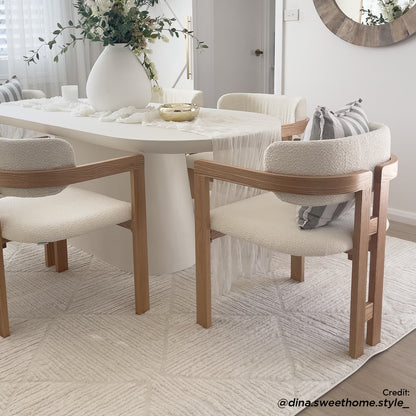 Maddi Boucle Hardwood Dining Chair in Cream White, features a boucle fabric, curved backrest, and natural oak frame, positioned around a white table with a table runner, vase, and decorative dish.