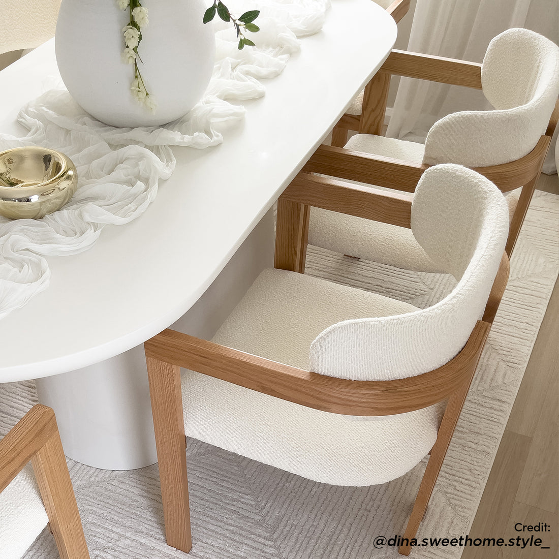 Maddi Boucle Hardwood Dining Chair in Cream White, featuring a curved backrest and upholstered in light boucle fabric, framed by warm wood, set around a modern white oval dining table.