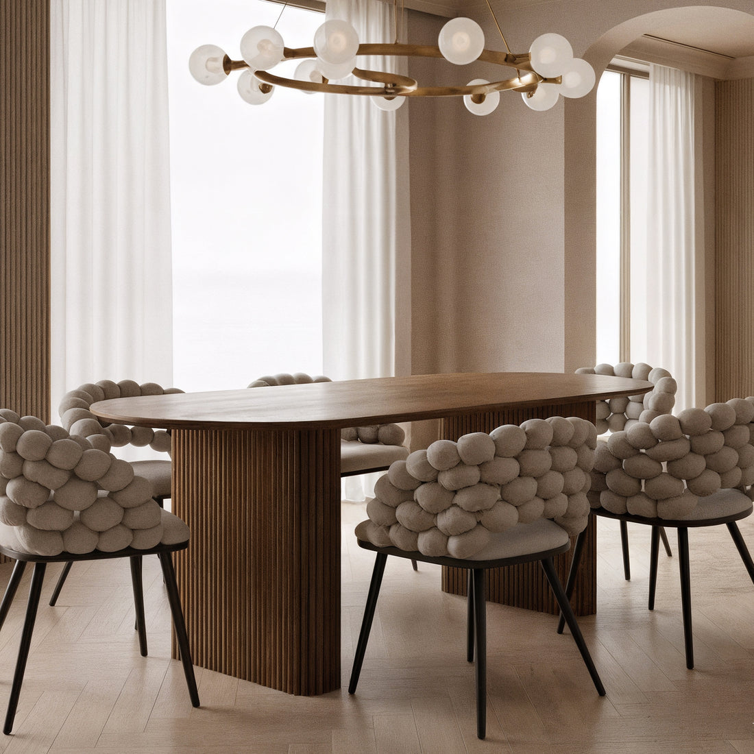 Marciana 8 Seater Fluted Oval Dining Table with a fluted wooden pedestal base in a softly lit dining room, surrounded by sculptural chairs featuring pebble-like cushions, under a modern brass-tone chandelier.