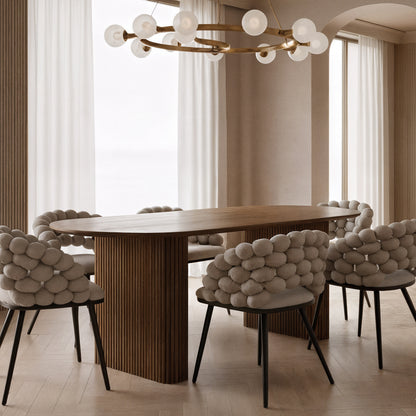Marciana 8 Seater Fluted Oval Dining Table with a fluted wooden pedestal base in a softly lit dining room, surrounded by sculptural chairs featuring pebble-like cushions, under a modern brass-tone chandelier.