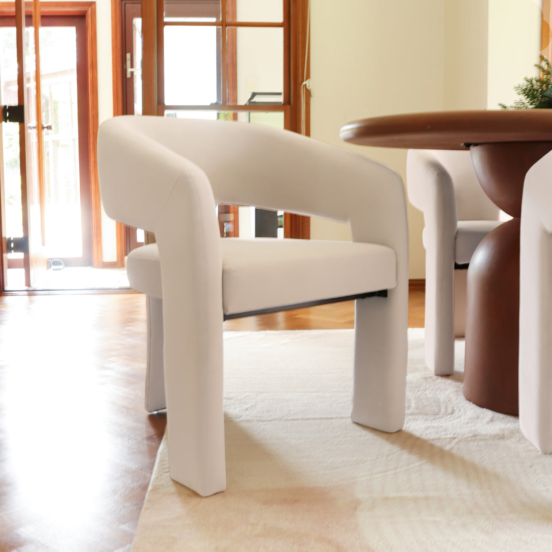 Mariah Velvet Upholstered Accent Chair with curved back in a bright, minimalist setting. Positioned by a round wooden table, it complements the light-colored rug, framed by tall glass doors.