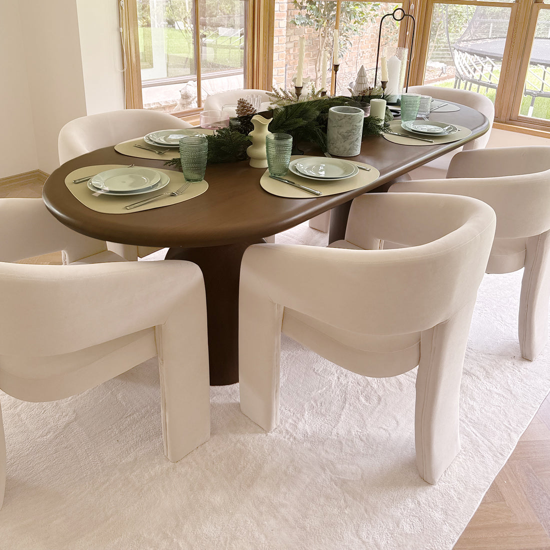 Dining nook with Mariah Velvet Upholstered Accent Chair in beige, featuring a sculptural wooden table, cream-upholstered chairs, seasonal evergreen centerpiece, and garden view through large windows.