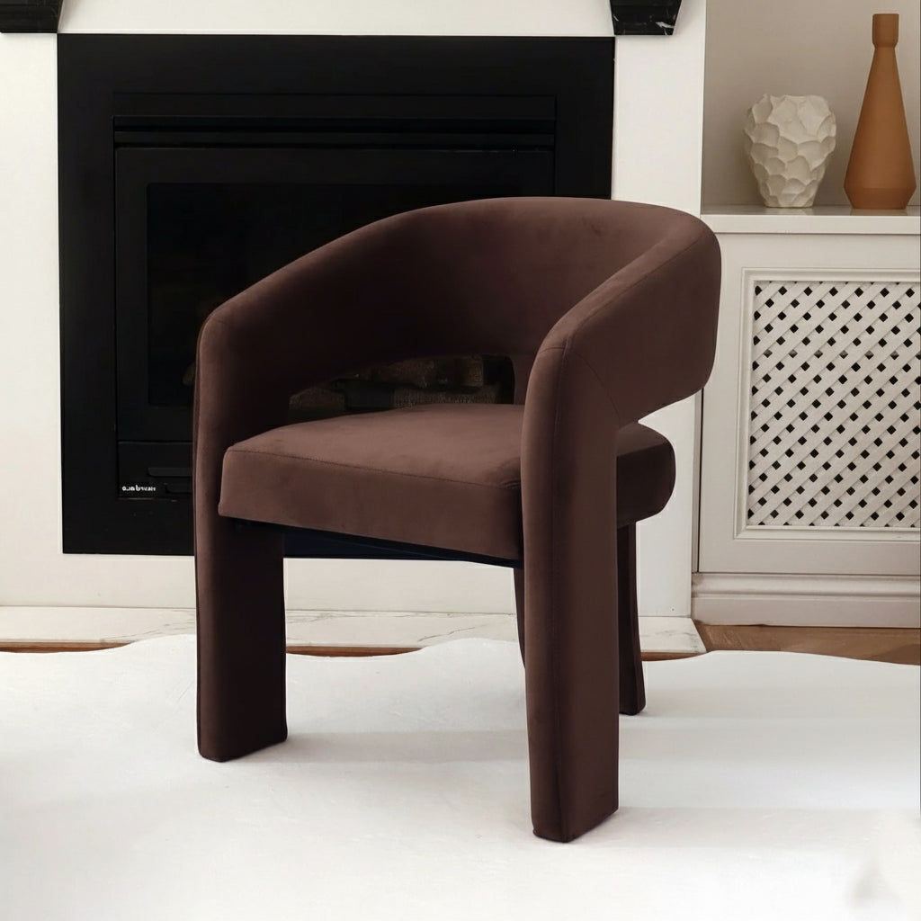 Mariah 3 Legged Velvet Accent Chair in Mocha Brown, showcased on a white rug before a black-trimmed fireplace, next to a white lattice cabinet adorned with two decorative vases.