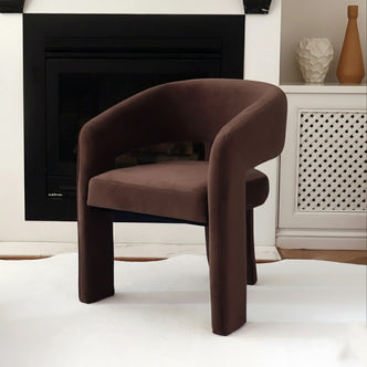 Mariah 3 Legged Velvet Accent Chair in Mocha Brown, showcased on a white rug before a black-trimmed fireplace, next to a white lattice cabinet adorned with two decorative vases.