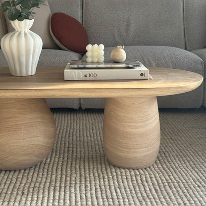 Mason 100cm Pillar Concrete Coffee Table in Beige, with a sculptural oval top and rounded legs, placed on a woven rug, adorned with decorative items including a vase and a marble tray.