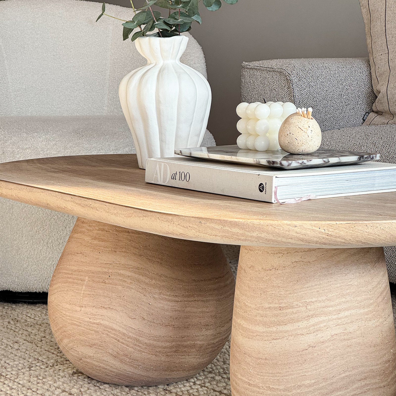 Mason 100cm Pillar Concrete Coffee Table in Beige with a sculptural vase, eucalyptus sprigs, AD at 100 book, marble tray, bubble candle, and stone match vessel atop, exuding minimalist elegance.