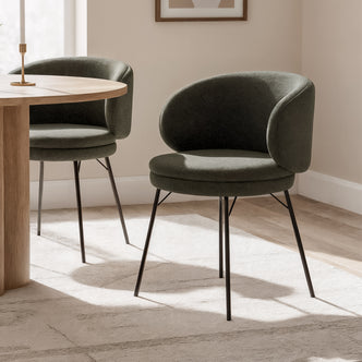 Meily Boucle Dining Chair Set of 2 in Green, featuring olive-green fabric on curved backs and seats, slender black legs, placed beside a wooden table in a sunlit, minimalist dining nook.