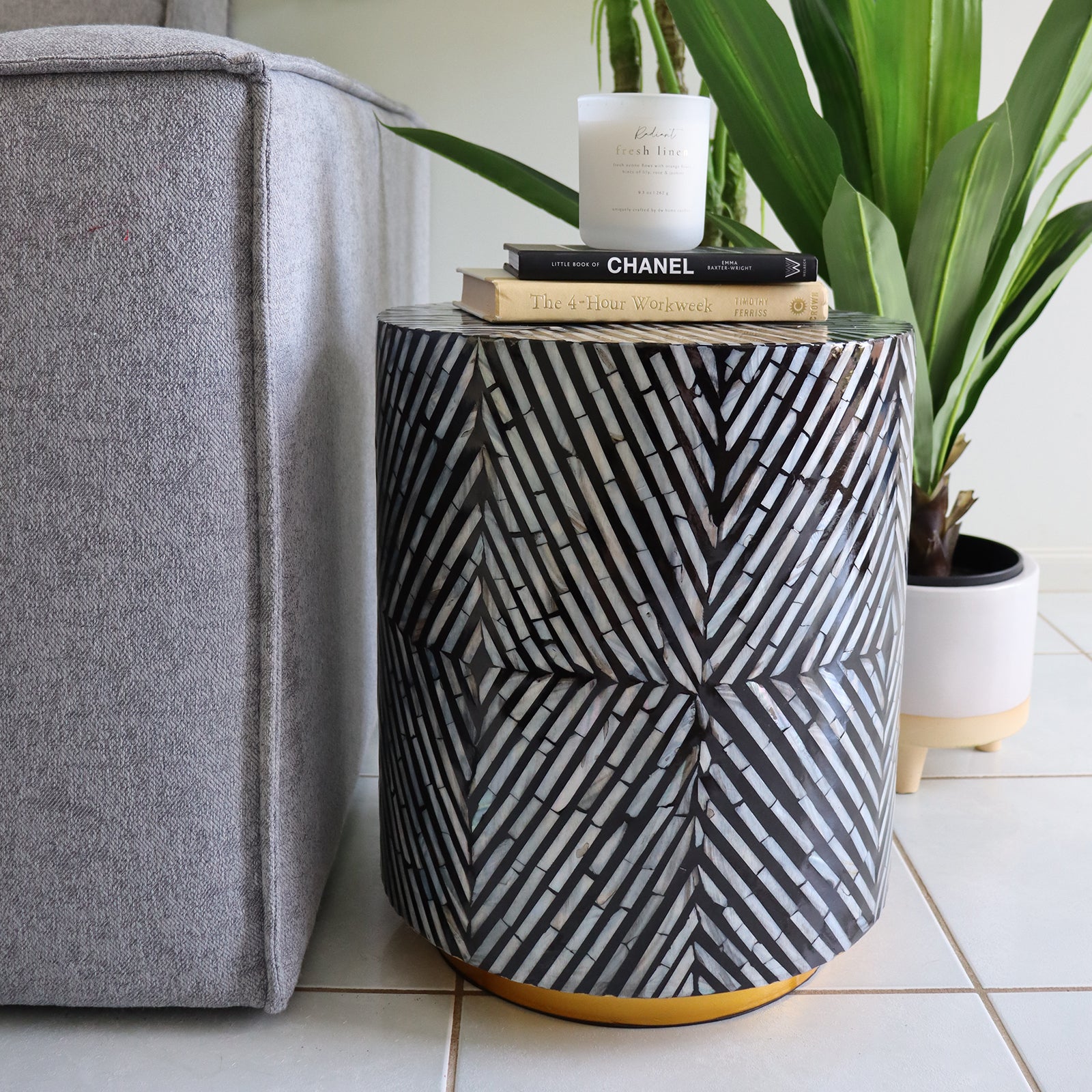 Merry 35cm Mother of Pearl Inlay Side Table in Ivory & Black beside a gray sofa, adorned with books and a candle, showcasing its elegant herringbone-style mosaic pattern.