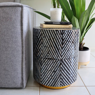 Merry 35cm Mother of Pearl Inlay Side Table in Ivory & Black beside a gray sofa, adorned with books and a candle, showcasing its elegant herringbone-style mosaic pattern.