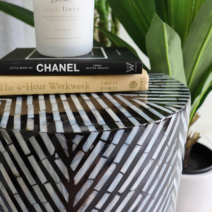 Merry 35cm Mother of Pearl Inlay Side Table in Ivory & Black featuring a geometric pattern, topped with two books and a frosted candle, framed by lush green plant leaves.