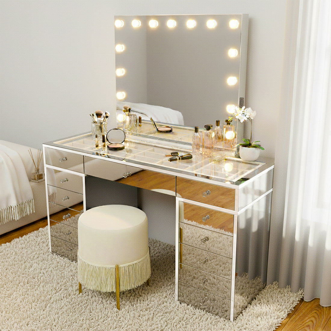 Monroe Hollywood Makeup Dressing Table in Silver features a large LED bulb mirror, glass-top with mirrored drawers, neat makeup arrangement, potted orchid, and a fringed stool on a plush rug.