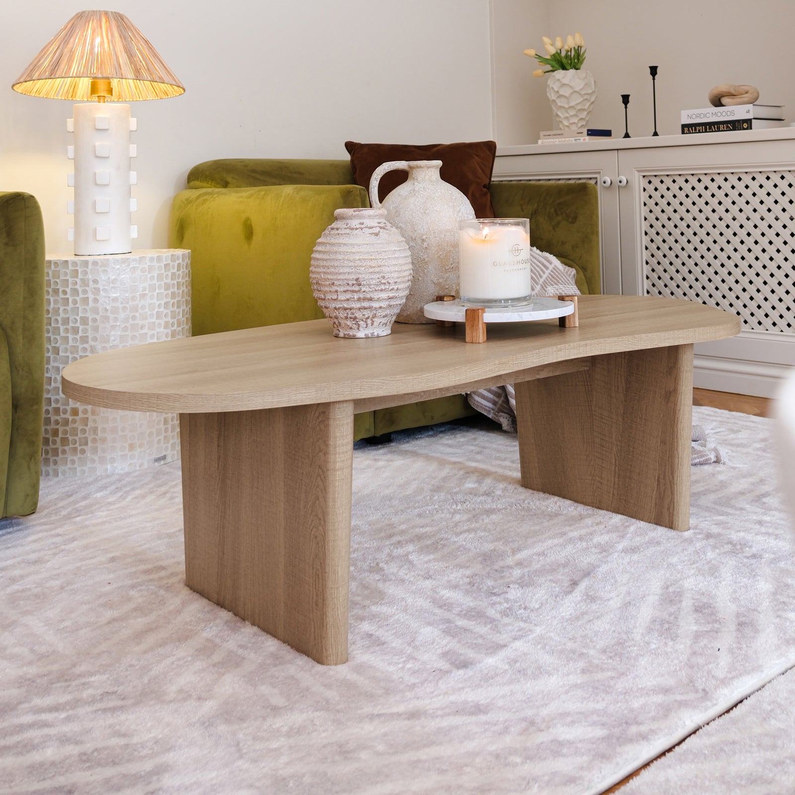 Nicole 120cm Oval Coffee Table in Natural, featuring an organic shape with soft curves and broad legs, adorned with two ceramic vases and a lit candle on a marble tray.