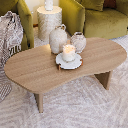 Nicole 120cm Oval Coffee Table in Natural features an organic kidney shape, natural wood finish, and minimalist design, adorned with rustic ceramic vases and a lit candle on a light patterned rug.